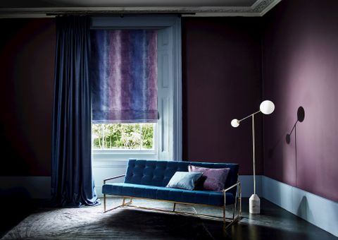 Living room with velvet sofa and single window dressed with curtains layered over a Roman blind