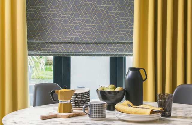 A table set for breakfast sits in front of a window featuring bright yellow blinds and a grey Roman blind that has a geometrical yellow design on it. The yellow of the curtains is echoed in the choice of yellow kitchen accessories