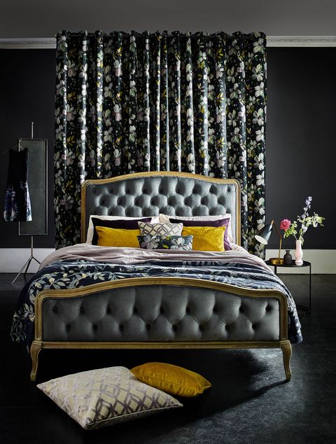 Dark painted bedroom with floral curtains behind a double bed with mustard, purple and pink pillows