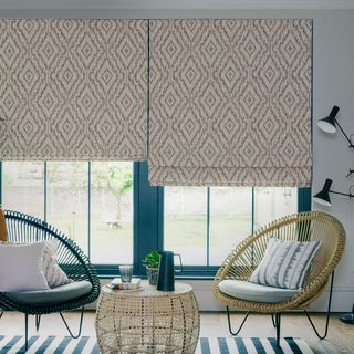 Cream patterned roman blinds in a white room that features large green plants. There are two wicker chairs with cushions in front of the window.