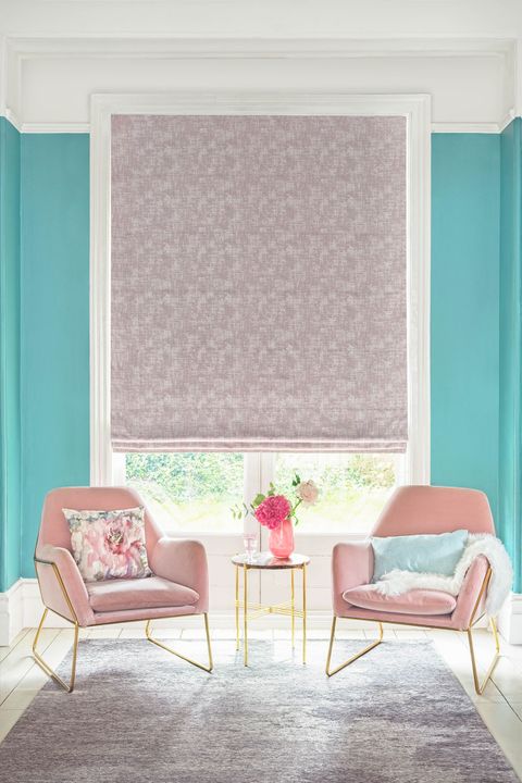 textured shimmering purple Roman blinds in living room. Light blue and printed cushions have been placed on two blush pink arm chairs with gold legs in the room