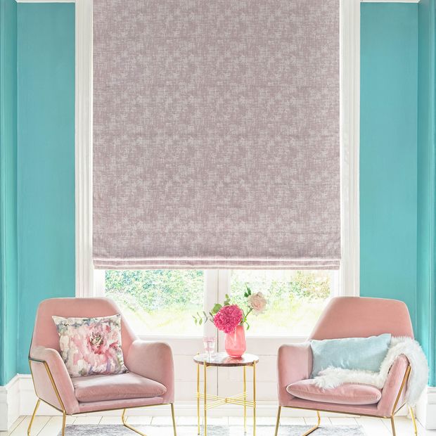 textured shimmering purple Roman blinds in living room. Light blue and printed cushions have been placed on two blush pink arm chairs with gold legs in the room
