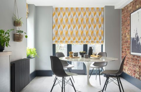 This grey and white room features a dining room table with four chairs. The long windows behind the table are covered with a Roman blind featuring a grey and yellow geometric diamond shaped design 