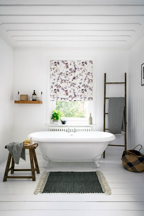In this white bathroom  window behind the white bath is dressed with white roman blinds printed with pink and purple flowers.