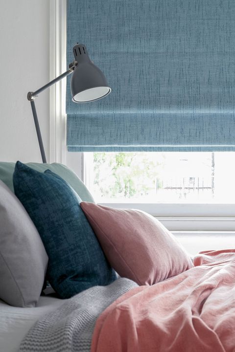A window featuring a sky blue Roman blind is behind a bed piled with blue and pink cushions and a throw