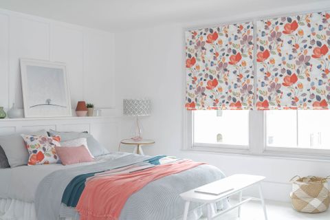  This white bed room features a double  bed piled with grey, white, floral and pink cushions and pink, blue and grey throws. A large window in the room is dressed with warm toned floral print on white background roman blinds.