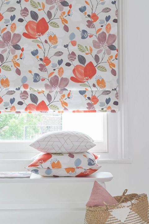 Close up of warm toned floral printed roman blinds in a bed room window. Matching floral and patterned cushions have been placed on white table in the room.