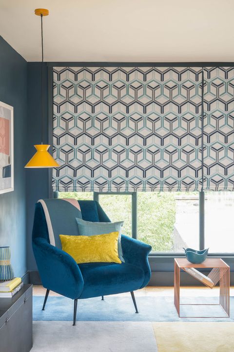 Close up of blue roman blinds featuring geometric shapes in teal and blue color on cream background. Light blue and mustard cushions have been placed on teal arm chair in the room.