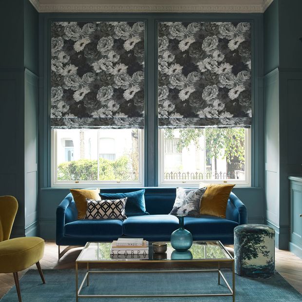 A blue couch rests in front of a window that is covered by a black floral Roman blind. The room is painted a deep forest green