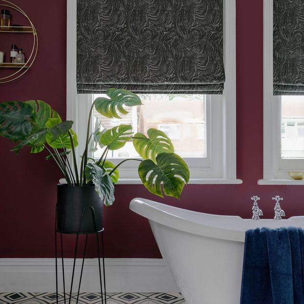 A deep wine bathroom with white bath and windows dressed with silver and grey marble effect roman blinds.
