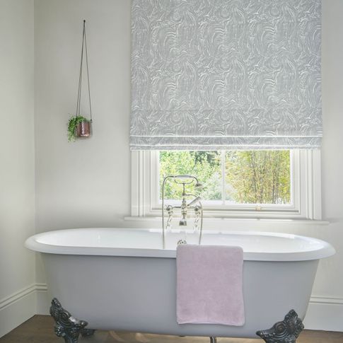 Cream bathroom with a white bath and white windows dressed with silver marble effect roman blinds.