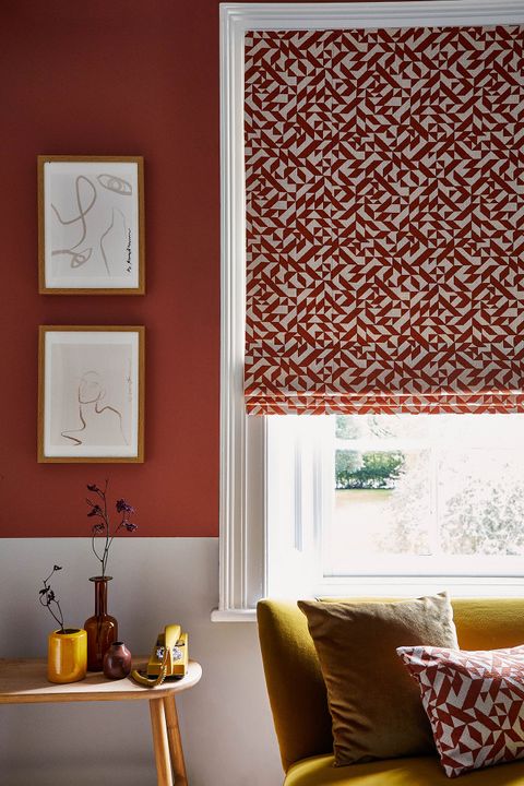 Corner view of a red and white room with a bright yellow couch that sits under the window. The window is covered with a Roman blind that features a red and white linear design