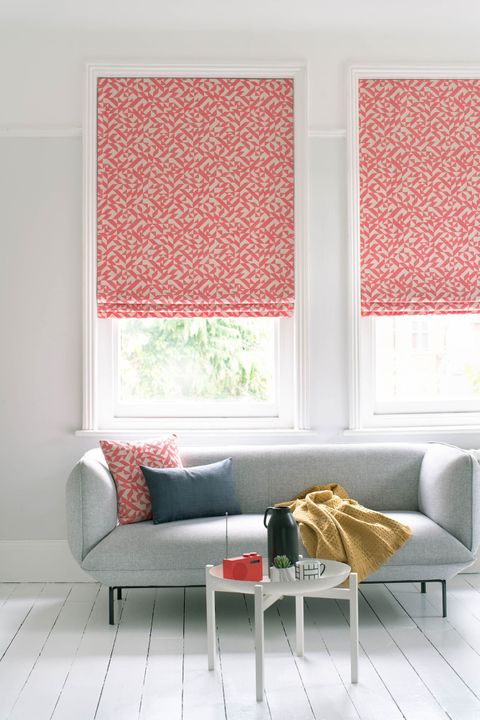 Windows of room white room are dressed with roman blinds featuring linear print in bright red and white color. Cushion matching to roman blinds have been placed on grey sofa.