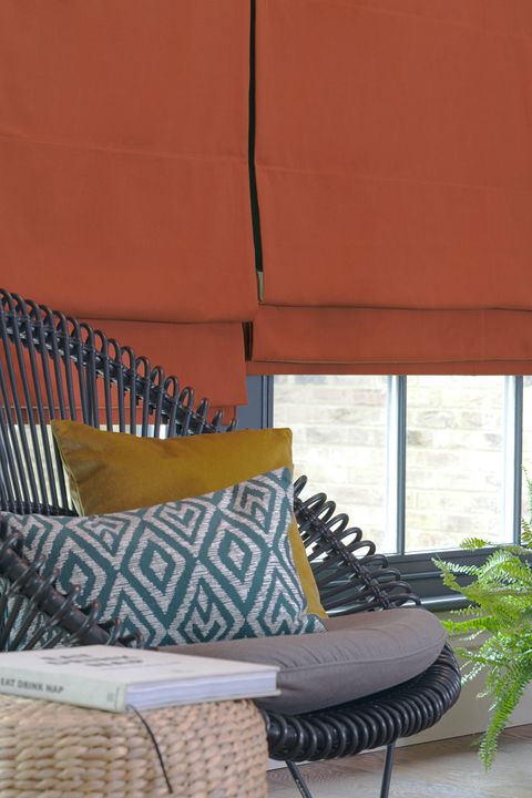 View of a window dressed with orange plain roman blinds. Blue patterned cushion and mustard plain cushion have been placed on a wicker chair resting in front of window.