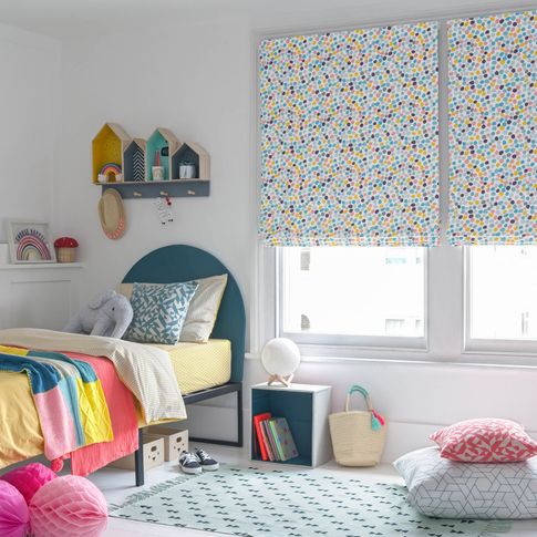 Children bedroom window dressed with roman blinds featuring multi-coloured dot pattern on a white background. Geometric printed cushions have been placed on  blue bed and rug in the room.
