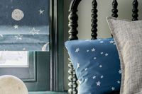 View of blue roman blinds printed with stars and moon hanging on a kids bedroom window. Bed is dressed with blue cushion printed with white stars.