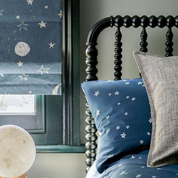View of blue roman blinds printed with stars and moon hanging on a kids bedroom window. Bed is dressed with blue cushion printed with white stars.