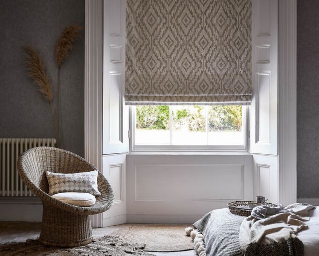  A patterned Roman blind hangs down over the window. A wicker chair has been placed nearby with cushions resting in the seat