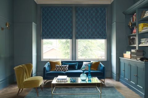 A blue room features a large window covered by a blue Roman blind that has a diamond shaped pattern. It hangs behind a blue sofa and a yellow velvet chair