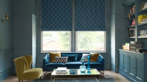 A blue room features a large window covered by a blue Roman blind that has a diamond shaped pattern. It hangs behind a blue sofa and a yellow velvet chair