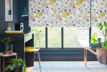 A multi-coloured floral Roman blind hangs neatly in front of a window on a petrol blue wall. Plants are dotted around the room and there is a desk to the left of the window. 