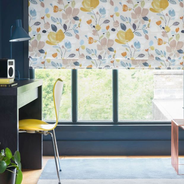 A multi-coloured floral Roman blind hangs neatly in front of a window on a petrol blue wall. Plants are dotted around the room and there is a desk to the left of the window. 
