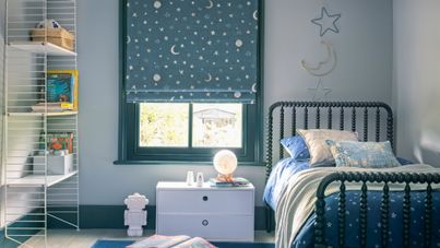 A child's bedroom with a blue roman blind featuring stars and moons. A star and moon decoration hangs on the right above the bed.