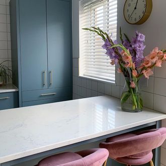 White wooden blinds hanging on window of kitchen. White breakfast table along with two pink velvet chairs has been placed near window