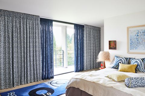 In the view white and blue retro print, plain unlined blue curtains are dressed on the sliding doors of bedroom. White and blue zebra printed cushions and citrine color plain cushion has been placed on the bed.
