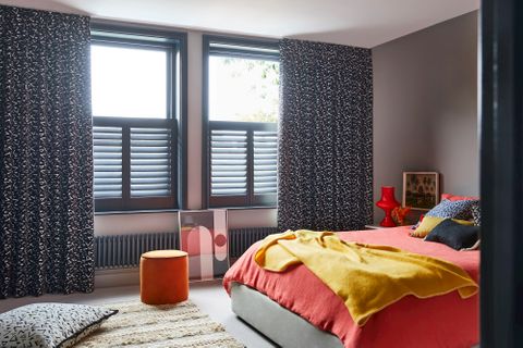 Black and white retro print curtains over grey cafe style shutters in a bedroom. Blue leopard cushion and Citrine plain cushion are placed on orange duvet on bed. White retro print cushion is resting on floor