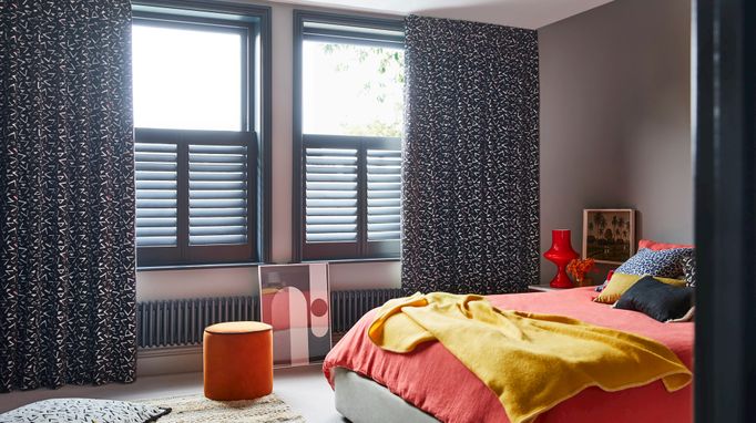 Black and white retro print curtains over grey cafe style shutters in a bedroom. Blue leopard cushion and Citrine plain cushion are placed on orange duvet on bed. White retro print cushion is resting on floor