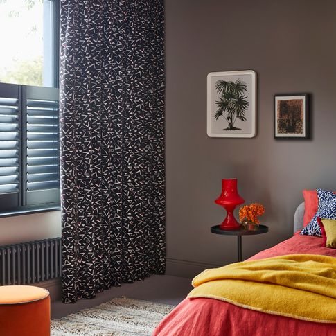 Corner view of bedroom where Black and white retro print curtains over grey cafe style shutters on windows. Blue leopard cushion and Citrine plain cushion are placed on orange duvet on bed. 