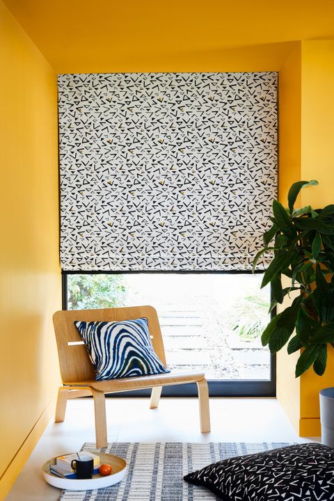 White roman blind featuring triangles in retro print hanging on window of yellow painted living room. Black retro traingle printed cushion is placed on the floor.