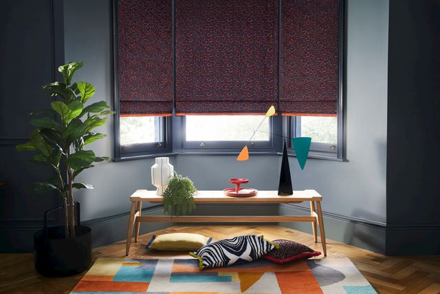 Dark grey roman blinds featuring bright orange boho inspired embroidery dressed on windows in living room. Zebra, boho print and plain cushions have been placed under table in the room.