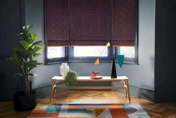Dark  grey roman blinds featuring bright orange boho inspired embroidery dressed on windows in living room. Blinds are decorated with orange fringe. 