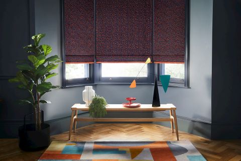 Dark  grey roman blinds featuring bright orange boho inspired embroidery dressed on windows in living room. Blinds are decorated with orange fringe. 