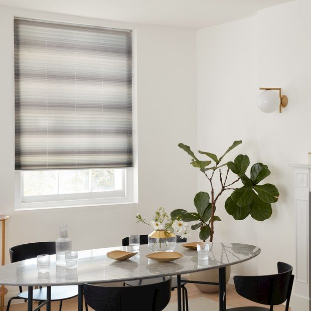 Side view mini stripe grey Pleated blinds from House Beautiful range hanging on single window in dining room. Grey dining table, four dark grey chairs in front and large green plant in corner.