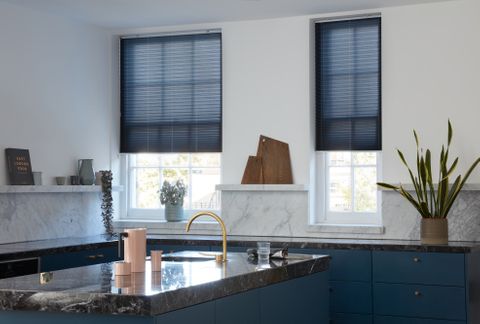 Blue Pleated blinds dressed on two kitchen windows. Blinds are matching colour to the cupboards and drawers of kitchen, with a peach jug accessory.
