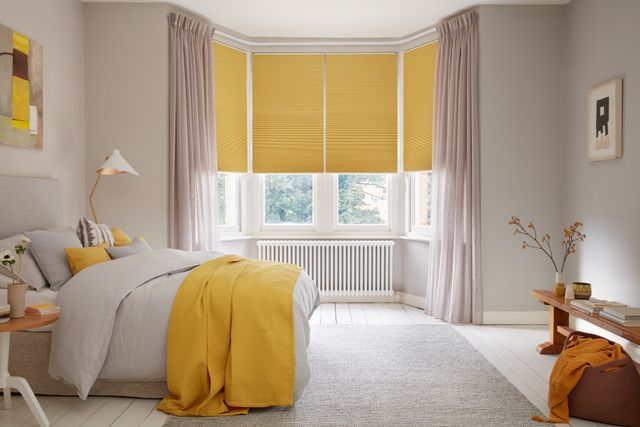 Mustard yellow blackout Pleated blinds dressed on windows in bedroom. Light pink Voile curtains are hanging over the blinds. Matching mustard and grey cushions have been placed on the bed.