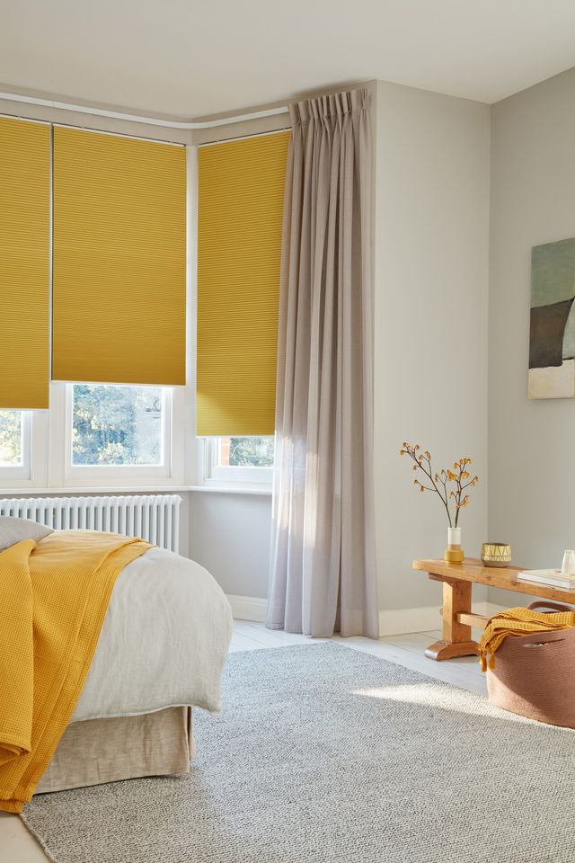 Corner view of bedroom where mustard blackout Pleated blinds dressed on windows. Almond Voile curtains are hanging over the blinds. Matching mustard and grey cushions have been placed on the bed.