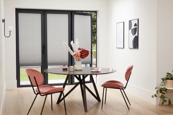 Grey Micro Pleat Pleated blinds dressed on open bi-fold doors of dining room. A round wooden table and peach/pink chairs are in the room.