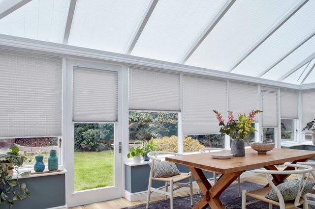 Grey motorised Pleated blinds dressed on windows of large conservatory. A large garden table along with chairs have been placed in conservatory.