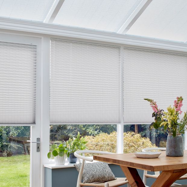 Grey motorised Pleated blinds dressed on windows of conservatory. An informal wooden table and relaxed dining chair sits on a grey rug.