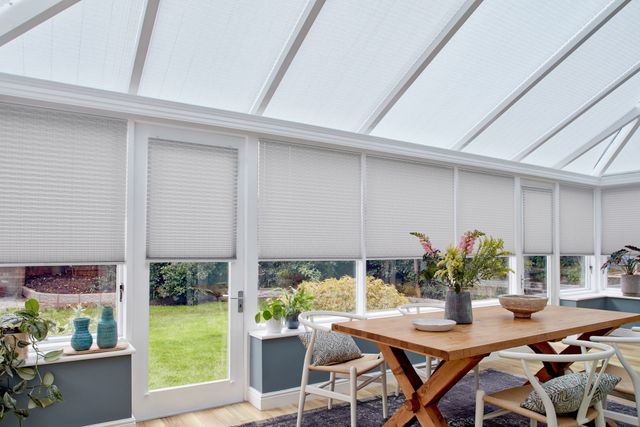 Grey motorised Pleated blinds dressed on windows of large conservatory. A large  table and chairs have been placed in conservatory, with several plants.