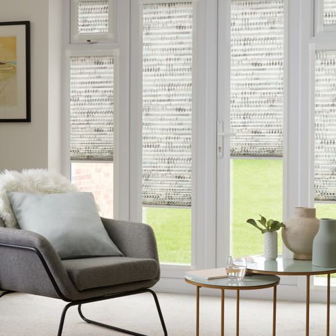 Duo natural Thermashade Perfect Fit Pleated blinds from House Beautiful range dressed on bi-fold doors, at varying drop heights. A grey upholstered chair and cushions are also in the room.