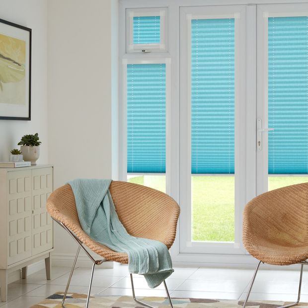 Bright blue Perfect Fit Pleated blinds on closed  bi-fold doors and small windows above doors. Two wicker chairs have been placed in the room, one with a pale blue throw draped across it.