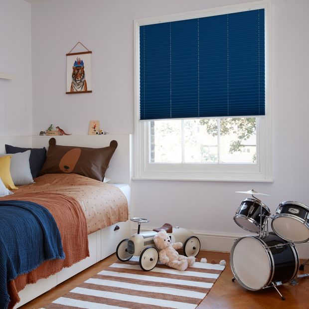 Dark blue Pleated blinds dressed on windows of kids' bedroom. Room has a single bed covered with brown cover, blue throws and cushions. A car, soft toy and drums are placed in the room.