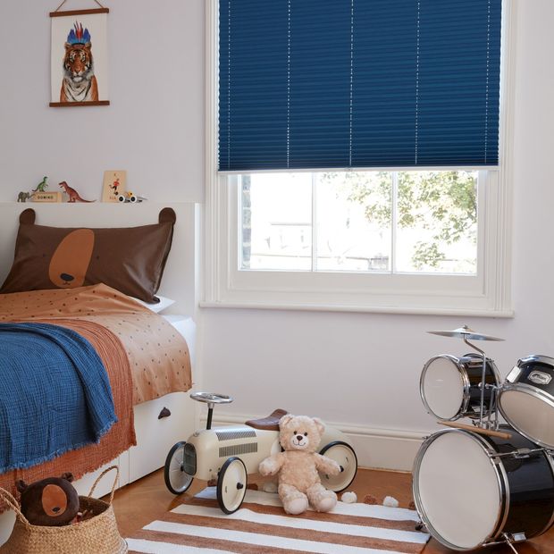 Dark blue Pleated blinds dressed on windows of kids' bedroom. A single bed with brown cover, blue throws and cushions sits alongside some toys and a small drum kit.