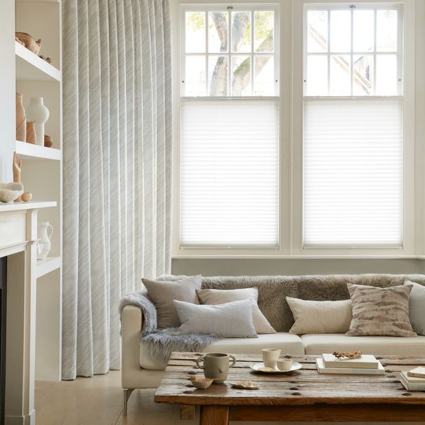 Corner view of a living room where white curtains featuring light grey pattern on curtains dressed over cafe style Pleated blinds on windows.