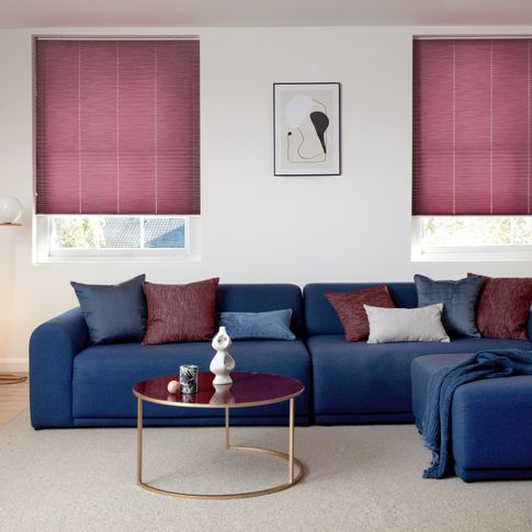 Maroon Pleated blinds dressed on windows of living room. Blue sofa decorated with maroon and white cushions have been placed in the room.